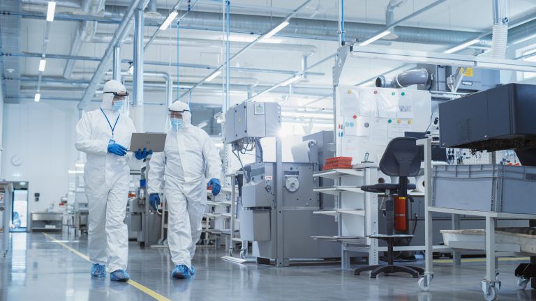 Two scientists in sterile coveralls walking on clean factory flooring, examining CNC machine settings